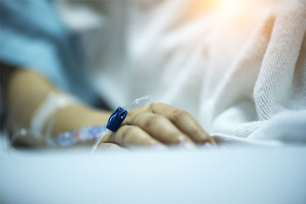 Paciente acostado con bata azul y cobija blanca con suero en la mano